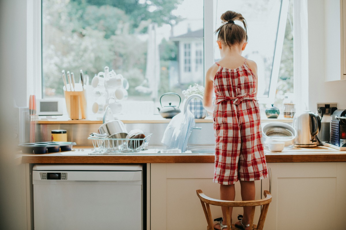 Little girl at the kitchen sink, a classic view out the window into the back yard. She's standing on a chair with an apron on, ready to help out.