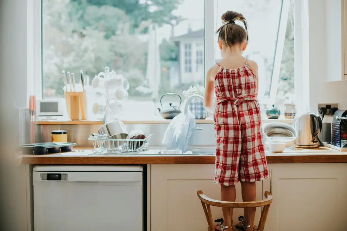 Little girl at the kitchen sink, a classic view out the window into the back yard. She's standing on a chair with an apron on, ready to help out.